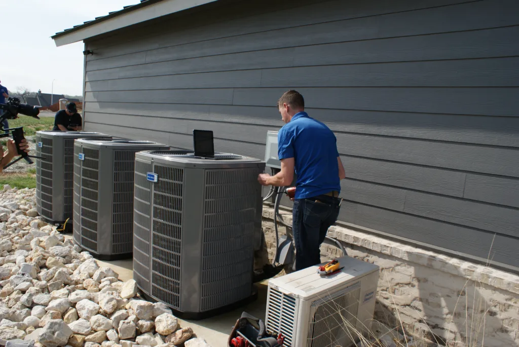 AC Replacement technician in a blue polo shirt is servicing outdoor air conditioning units next to a gray building.