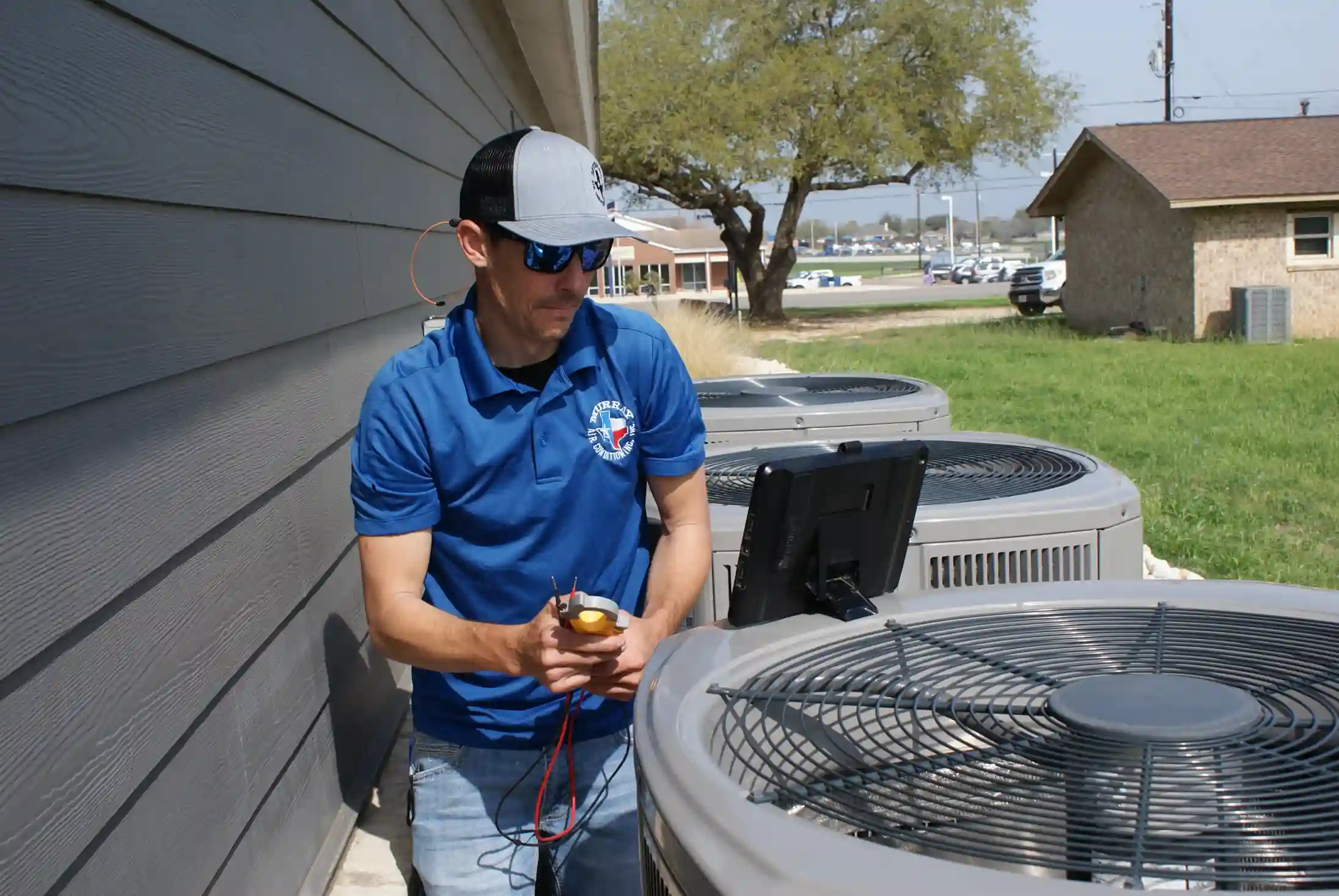 A technician in a blue polo shirt and a cap is working on an outdoor air conditioning unit.