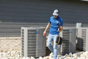A technician in a blue polo shirt and cap is walking away from outdoor air conditioning units.