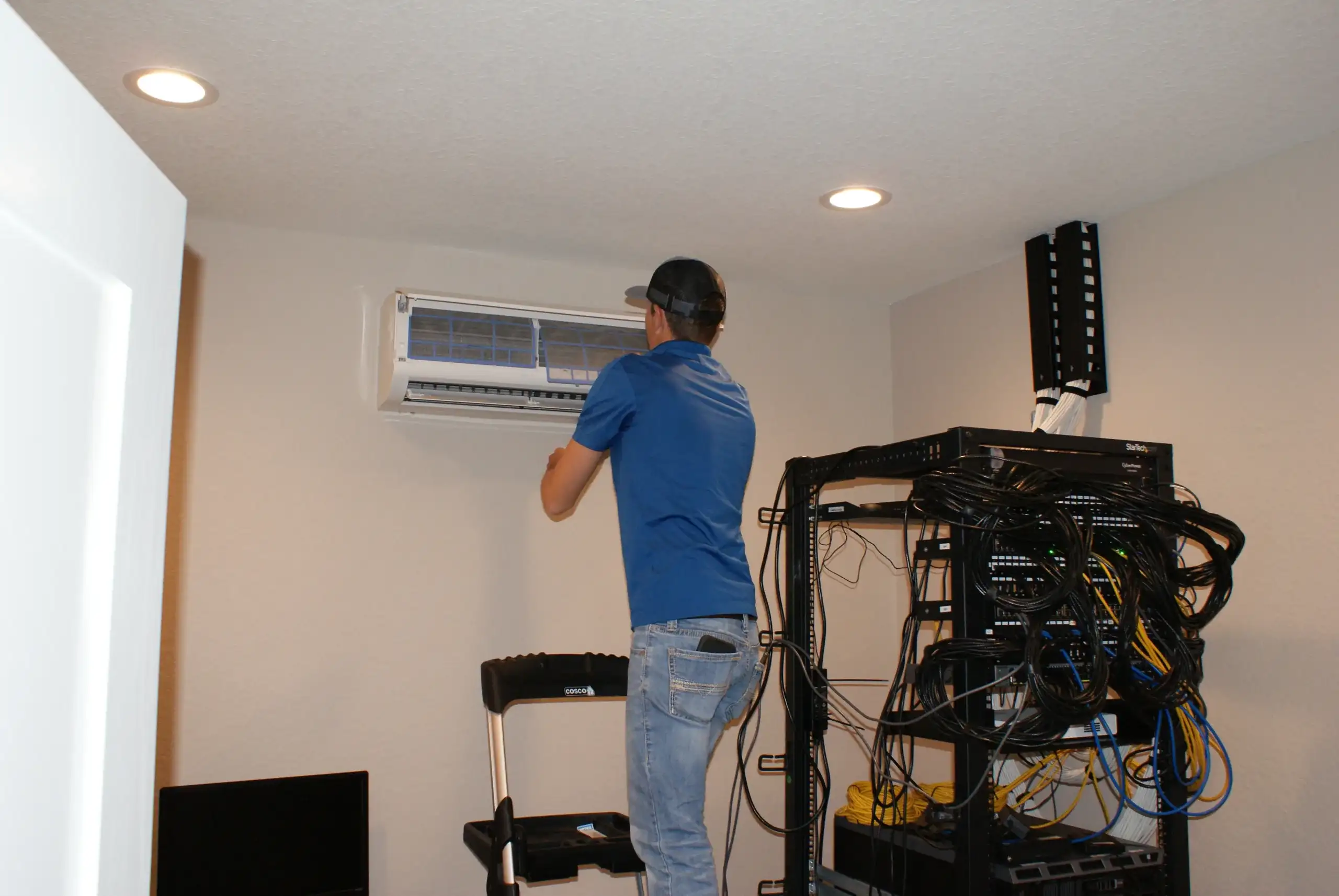 Office HVAC technician in a blue polo shirt and cap is standing on a step stool while working