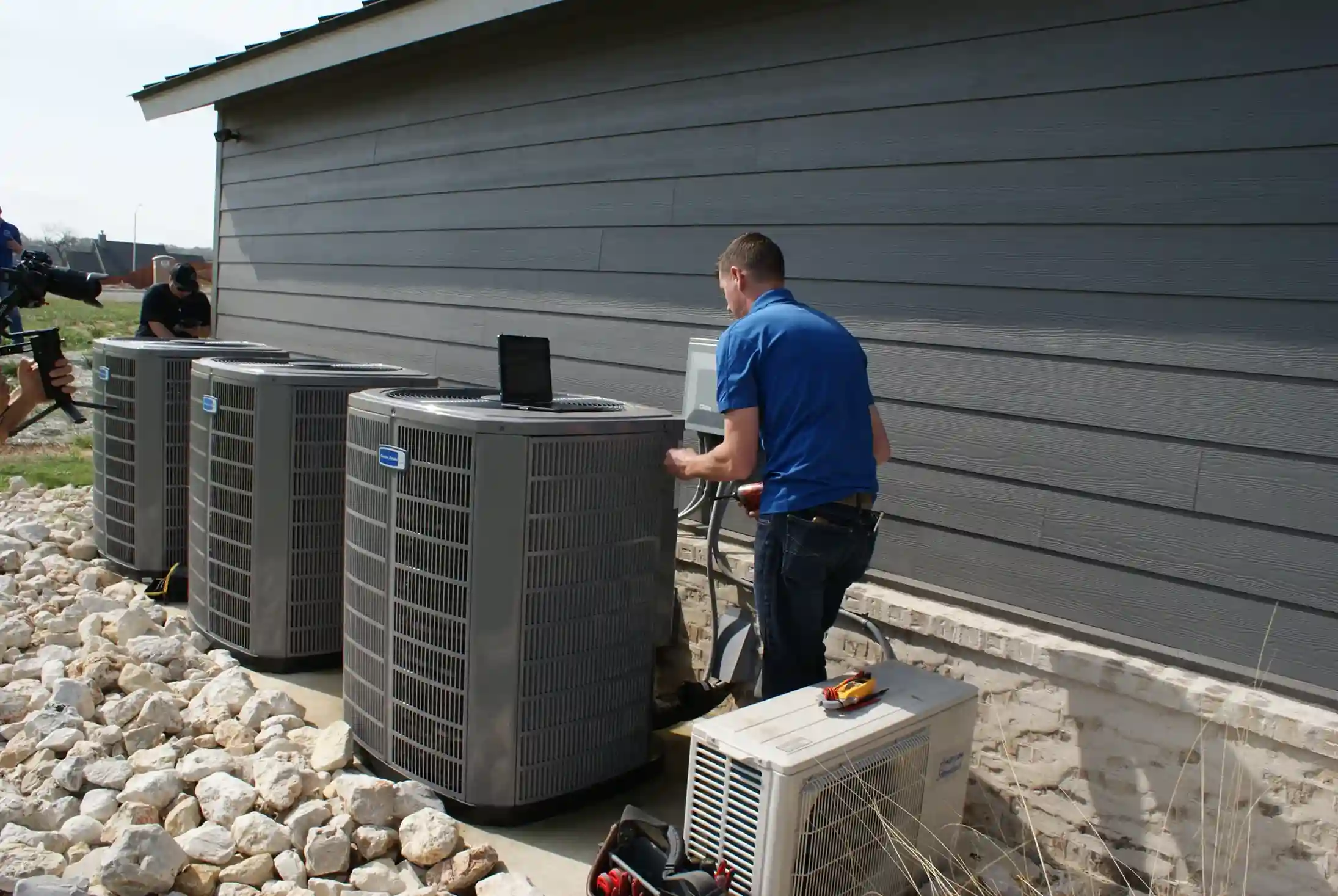 A technician in a blue polo shirt is servicing outdoor air conditioning units next to a gray building.