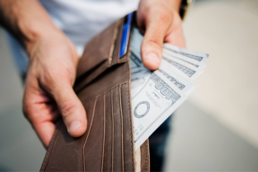 Person holding a brown wallet with multiple hundred-dollar bills visible.