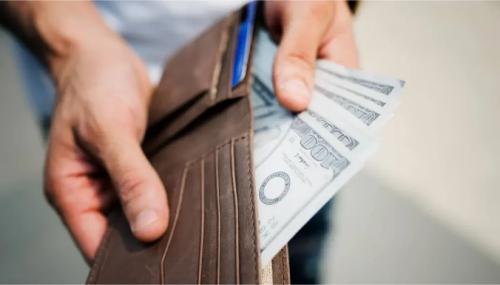 Person holding a brown wallet with multiple hundred-dollar bills visible.