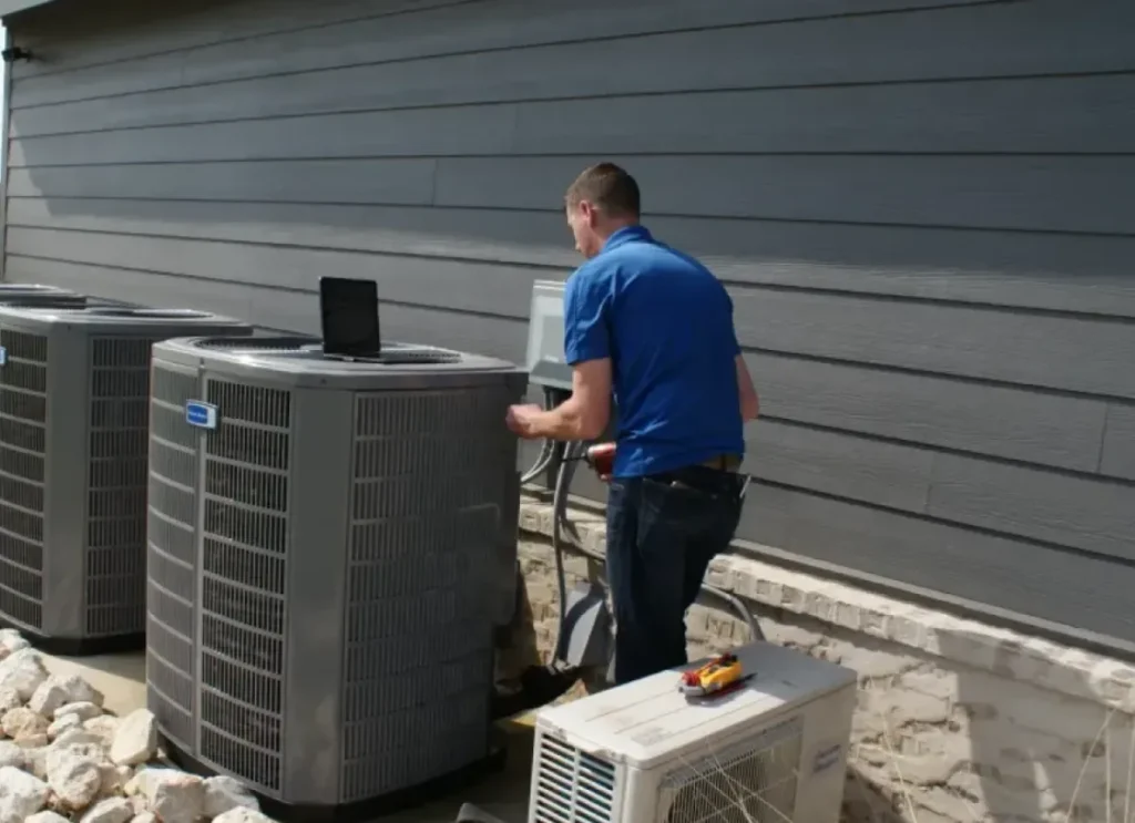 AC Replacement technician in a blue polo shirt is servicing outdoor air conditioning units next to a gray building.